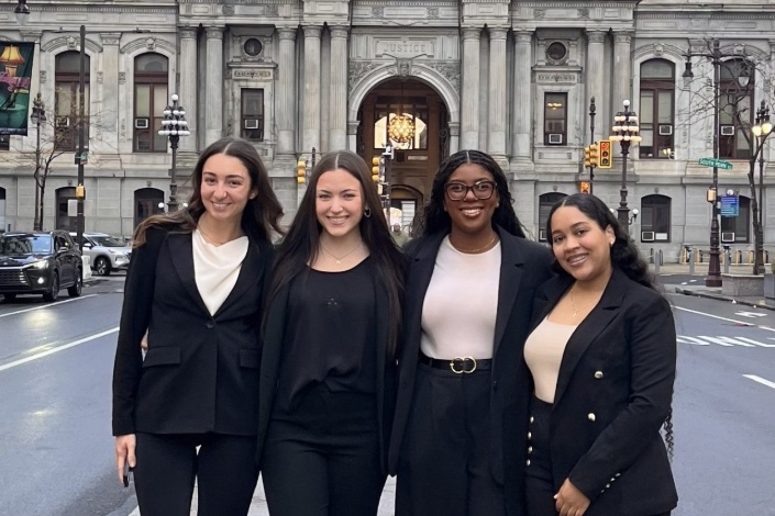 Four students pose together for a picture in front a big building in a city.