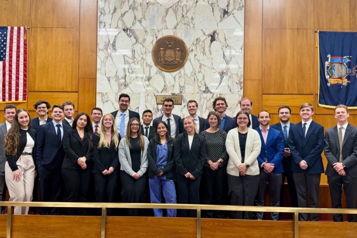 A large group of approximately two dozen young adults in professional attire stand in a courtroom, posing for a group photo. They are arranged in rows behind a wooden railing, with a judge’s bench and a marble wall behind them. An official seal is mounted on the wall at center, flanked by the U.S. flag on one side and a New York State flag on the other. The group appears formal and celebratory, as if marking a mock trial or academic legal event.