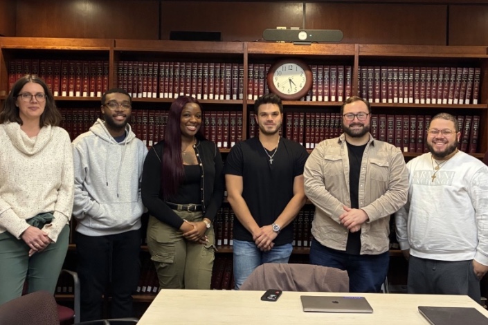 Seven people stand side by side in a conference room in front of floor-to-ceiling bookshelves filled with red-bound law books. A clock hangs above the group. The group is smiling and dressed in a mix of casual and business-casual clothing. A conference table with laptops is visible in the foreground.