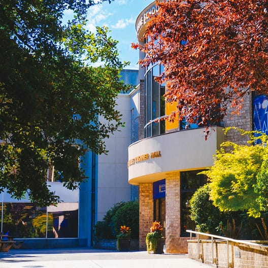 A modern building with large windows and a curved entrance is surrounded by lush green trees and red-leaved foliage on a sunny day. A blue sky with clouds is visible in the background.