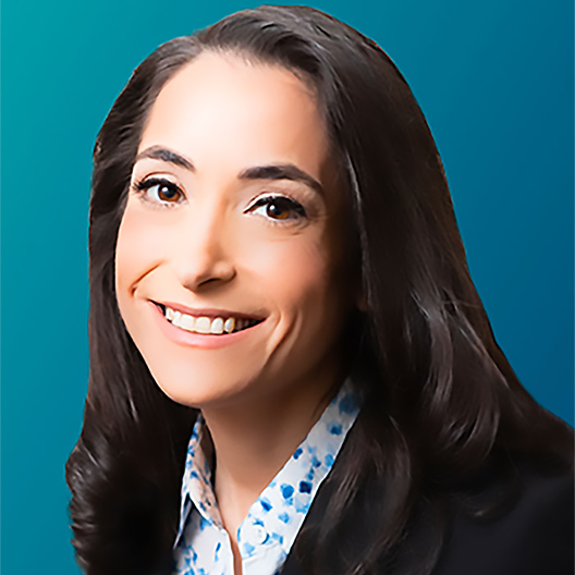 Ashira Pelman Ostrow, a smiling woman with long dark hair, wearing a patterned shirt and dark blazer, posed in front of a blue gradient background.
