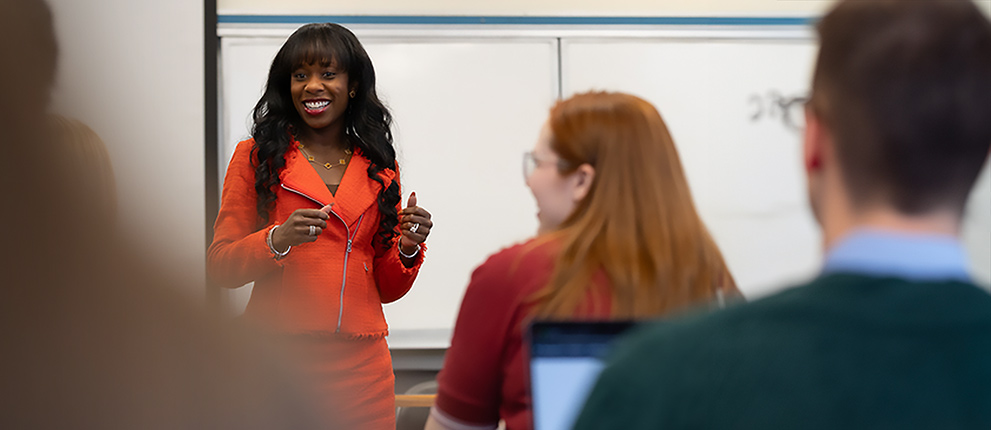 A woman in a bright red suit stands and speaks in front of a classroom, engaging with students who are listening and taking notes. A whiteboard is visible behind her.