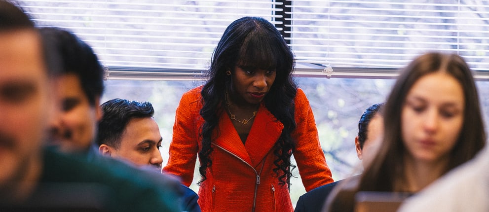 A woman in a bright orange jacket stands and looks down at a group of seated people, who appear to be engaged in discussion or work, in a classroom or office with large windows in the background.