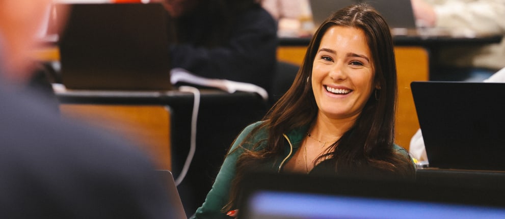 A woman with long dark hair smiles while sitting in a classroom with laptops and other students in the background.