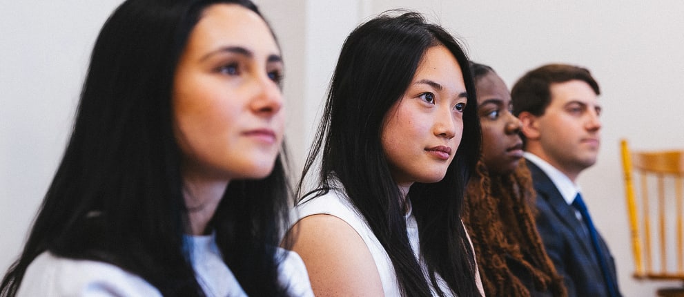 Four people, two women in the foreground and one woman and one man in the background, sit in a row inside a room, attentively looking toward something outside the frame.