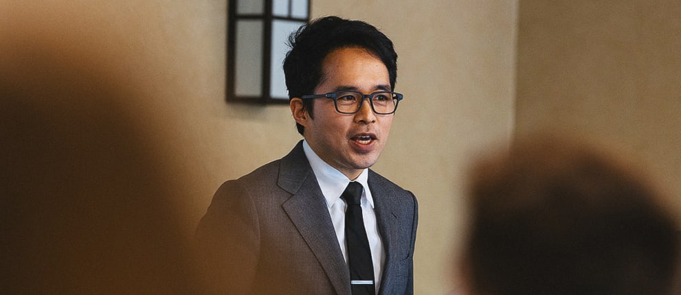 A man in a suit and glasses stands at the front of a room, speaking to an audience. The photo is taken from behind the audience, focusing on the speaker.