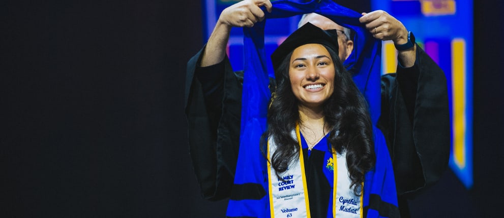 A smiling graduate in a cap and gown receives a blue hood on stage during a graduation ceremony. The background is dark, with some bright, colorful lights visible.