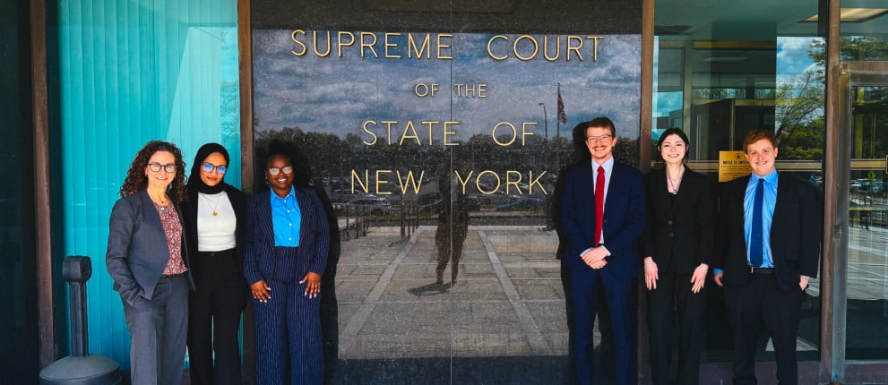Six people dressed in business attire stand smiling outside the Supreme Court of the State of New York, next to a reflective glass wall displaying the court’s name.