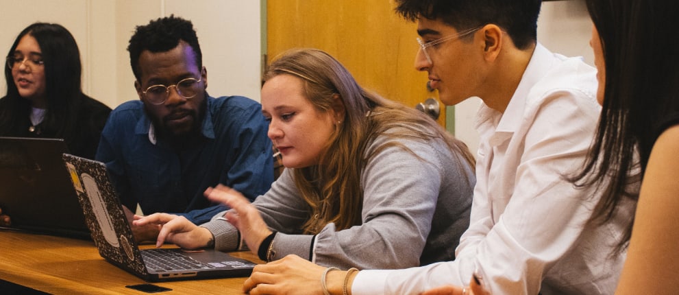 Five people are sitting around a table with laptops, engaged in discussion or collaborative work. Two people are closely looking at one laptop, while others observe or participate. The setting appears to be a meeting or study session.