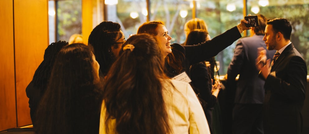 A group of people dressed in business attire gather indoors, with one woman holding up a phone to take a selfie. Warm lighting and large windows in the background create a lively, social atmosphere.