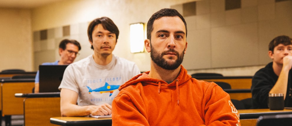 Three men sit in a mostly empty classroom. The man in front wears an orange hoodie and has his arms crossed. Two others sit behind him, one using a laptop. All are looking forward.