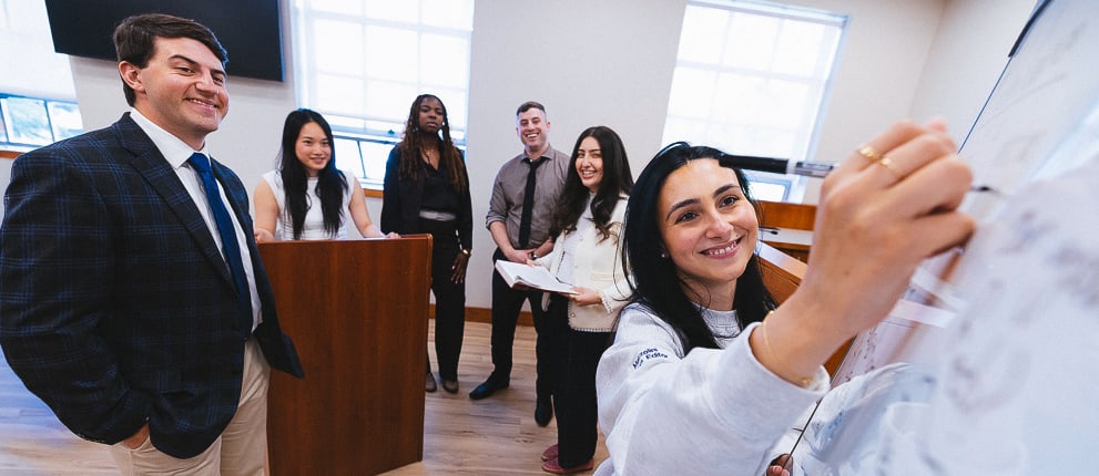 A group of six people in business attire stand in a bright room; one person smiles while writing on a whiteboard, and others stand behind a podium, smiling and looking at the camera.