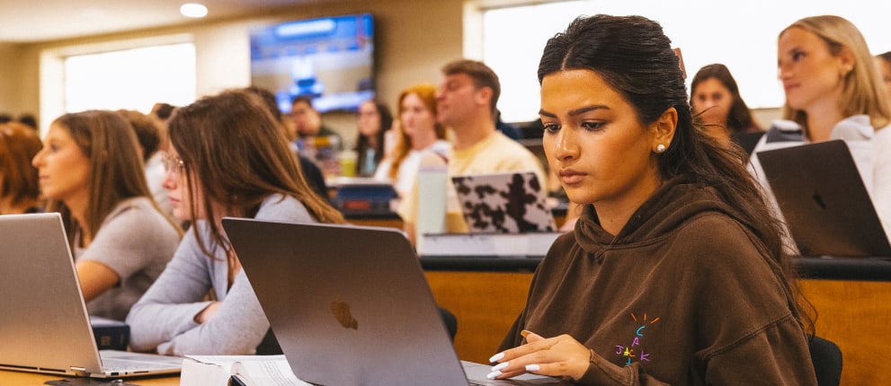 A focused student in a brown hoodie works on a laptop in a classroom, surrounded by other students using laptops and taking notes.