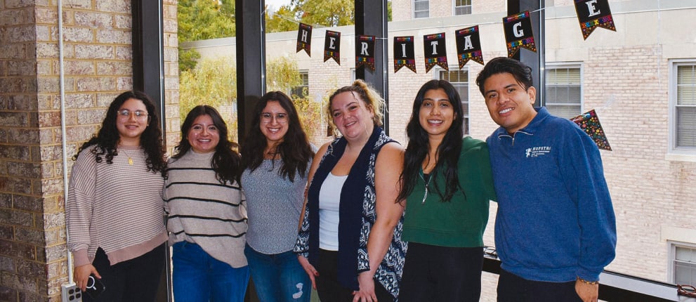 Six people stand indoors smiling at the camera, near large windows. Behind them, a “HERITAGE” banner is displayed. The scene appears casual and friendly, with natural light filling the space.