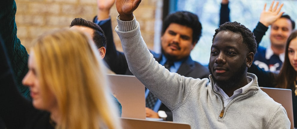 A group of diverse students sitting in a classroom, raising their hands to participate, with laptops open in front of them.