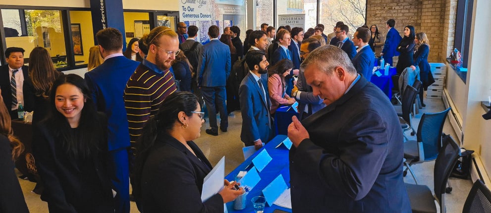 A large group of professionally dressed people interact at a busy indoor networking or career fair event, talking at tables and exchanging documents.