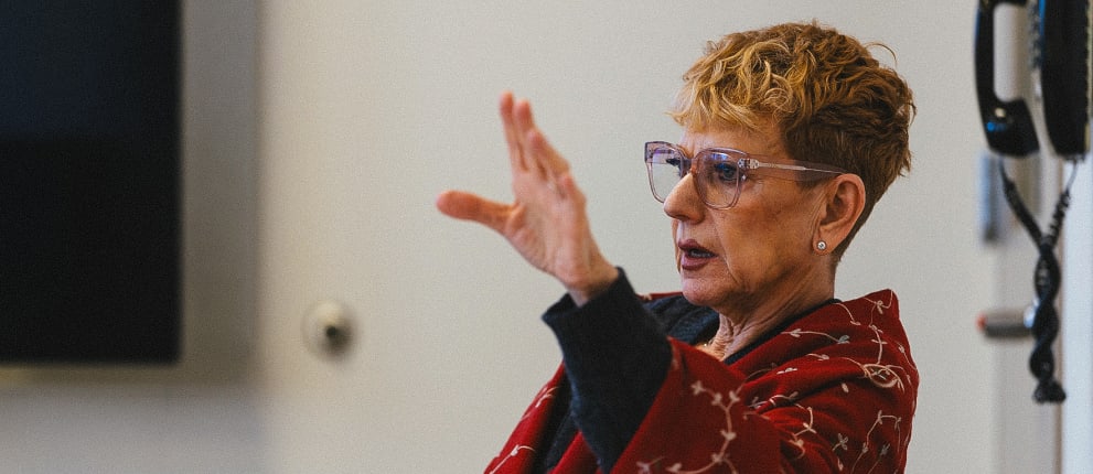A person with short curly hair and glasses, wearing a red patterned jacket, gestures with one hand raised while speaking in a modern indoor setting.