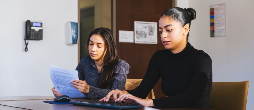 Two women sit at a desk in an office; one types on a keyboard while the other reads documents. There is a wall phone and posters in the background.