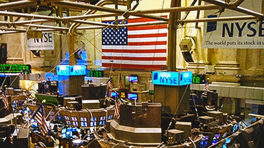 Numerous trading stations with computer monitors and NYSE signs fill a busy trading floor, with a large American flag displayed prominently on the wall in the background.