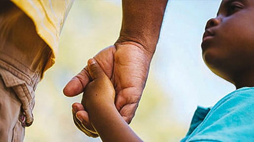 A child holds an adults hand while looking up, both wearing colorful clothing. The background is blurred, emphasizing their connection and the childs expression.