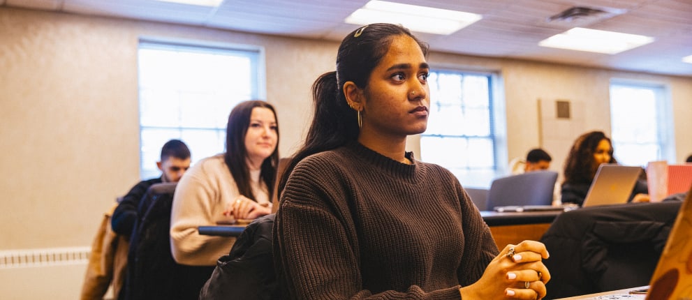 A young woman sits attentively in a classroom, hands clasped, with other students seated behind her. Bright windows let in natural light, and laptops and notebooks are visible on desks.