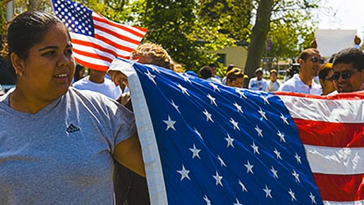 A woman holds a large American flag during an outdoor gathering with several people, some holding smaller flags, on a sunny day.