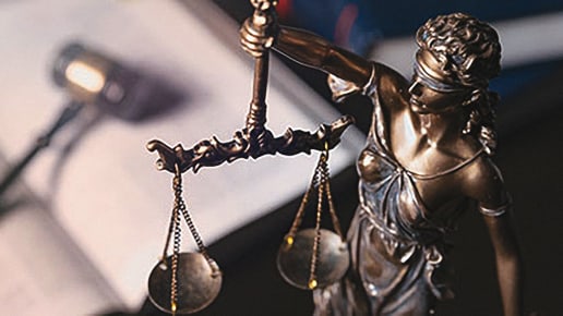 A close-up of a bronze Lady Justice statue holding balanced scales, with a blurred background featuring a microphone and papers, symbolizing law and justice.