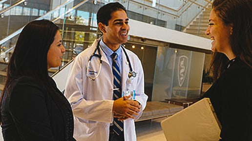 A doctor in a white coat speaks with two women in a modern building lobby. One woman holds a folder. They are smiling and engaged in conversation near a staircase and glass wall.