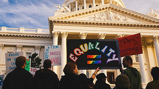 A group of people stand in front of a large government building holding colorful protest signs, including one that says “EQUALITY” with a rainbow equals sign. The sky is partly cloudy.