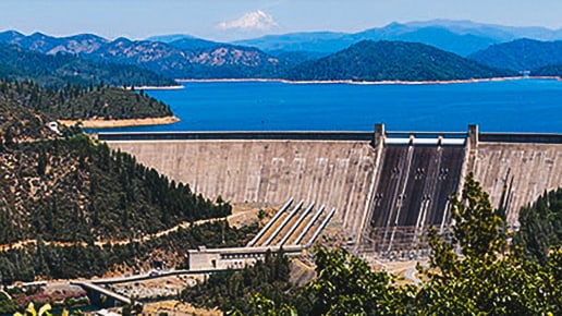 A large concrete dam holds back a blue reservoir surrounded by forested hills and distant mountains under a clear sky.