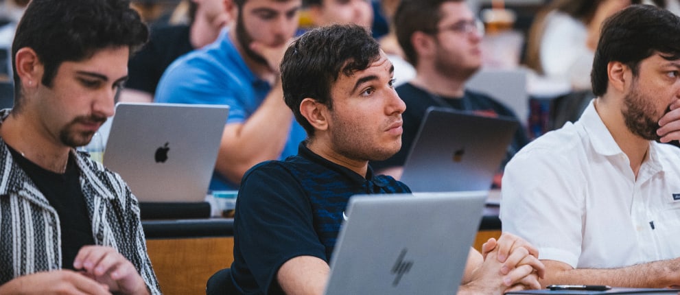 Students sit in a lecture hall, listening attentively. Some take notes and use laptops, while others focus on the speaker. The classroom is filled with rows of students engaged in learning.