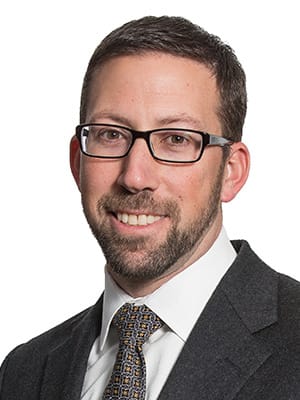 Photo of Christopher Caruso with short brown hair, beard, and glasses, wearing a dark suit, white shirt, and patterned tie, smiling at the camera against a plain white background.