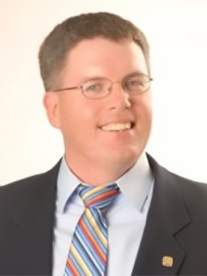 Photo of Michael G. Wagner wearing glasses, a dark suit jacket, a light blue shirt, and a colorful striped tie, smiling at the camera against a plain light background.