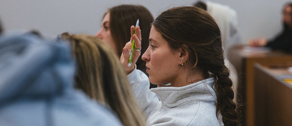 A student with a braid raises her hand in a classroom, holding a pen, while others sit and listen in the background.