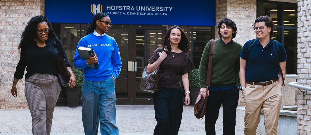 Five students walk and smile in front of the Hofstra University Maurice A. Deane School of Law building, holding bags and books, with the campus entrance sign visible in the background.