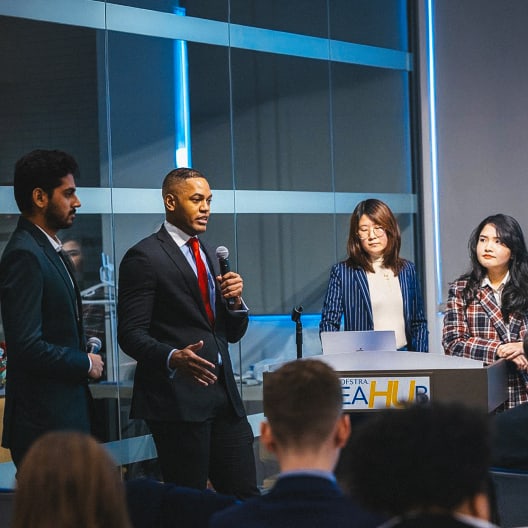 Four people in business attire stand and speak at the front of a room with an audience, participating in a panel discussion or presentation at Hofstra IdeaHub. A microphone and lectern are visible.