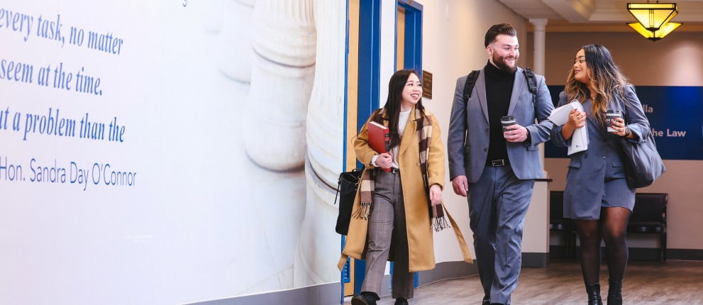Three professionally dressed young adults walk together in a hallway, smiling and holding coffee, books, and notebooks. A large wall with writing and a yellow emblem are visible in the background.