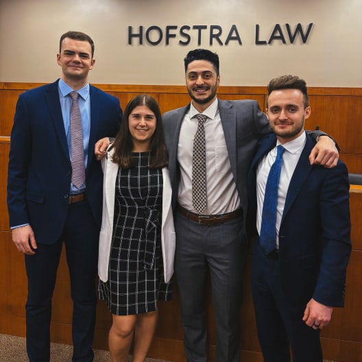 Four people dressed in business attire stand smiling with their arms around each other in front of a wall that says HOFSTRA LAW. They appear to be in a courtroom or formal setting.