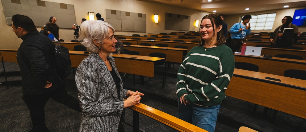 An older woman in a gray blazer speaks with a younger woman in a green striped sweater inside a mostly empty lecture hall, while several other people are gathered and talking in the background.