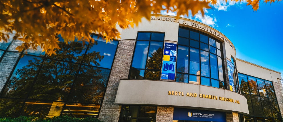 A modern building with large glass windows reflecting trees, labeled Maurice A. Deane School of Law and Seryl and Charles Kushner Hall, beneath orange autumn leaves.