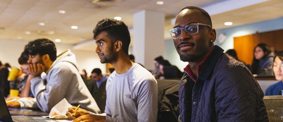 A group of students sits in a lecture hall, some taking notes and others listening attentively. One student in front, wearing glasses and a dark jacket, looks toward the camera and smiles.
