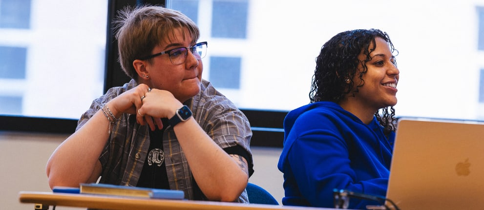Two students sit at a desk in a classroom. One wears glasses and looks to the side, while the other smiles and uses a laptop. Large windows with blurred city buildings are in the background.