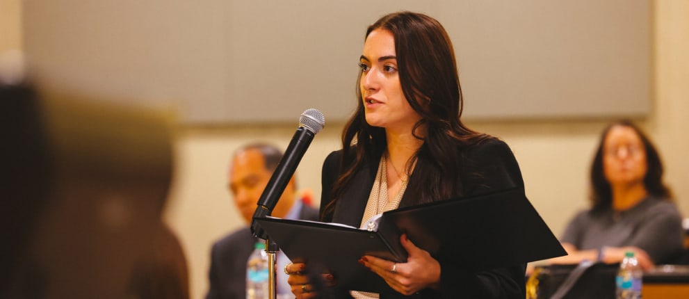 A woman in professional attire speaks into a microphone while holding a folder. Several people sit blurred in the background, suggesting a meeting or conference setting.