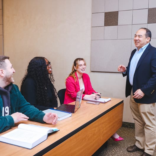 Three students sit at a classroom table with books and a laptop, smiling and engaging with a standing instructor who is speaking to them. The setting is bright and appears to be a modern classroom.