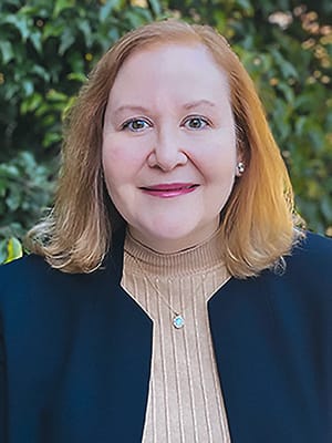 A photo of Lori Rowan with shoulder-length light brown hair smiles at the camera. She is wearing a beige turtleneck, a dark blazer, and a pendant necklace. Green leafy plants fill the blurred background.