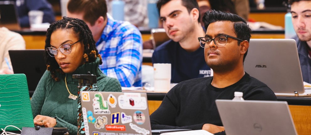 A diverse group of college students sit in a lecture hall, focused on the lesson. They have laptops open on their desks and appear to be listening attentively to the instructor.