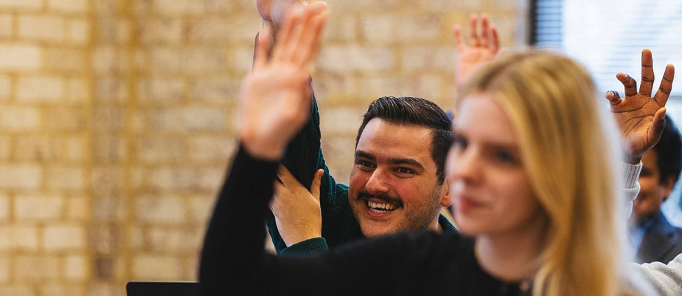 Several people in a classroom setting raise their hands, with a blurred woman in the foreground and a smiling man in focus behind her. The background features a brick wall and a window to the right.