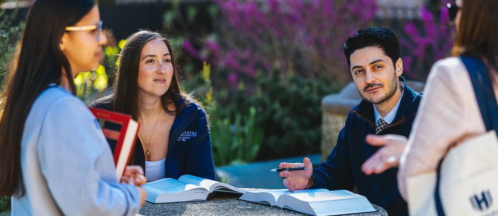 Three students sit at a round outdoor table with open books, talking and studying together. Another person stands nearby holding a laptop. Lush greenery and pink flowers are in the background.