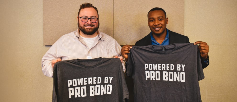 Two men smiling and holding up gray t-shirts that say Powered by Pro Bono in white letters, standing indoors against a neutral background.