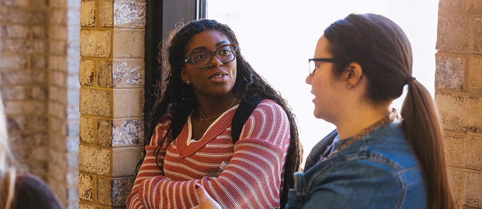 Four women stand indoors by a brick wall and window, engaged in conversation. One woman in a red striped sweater listens with arms crossed while another gestures as she speaks. The scene appears casual and friendly.